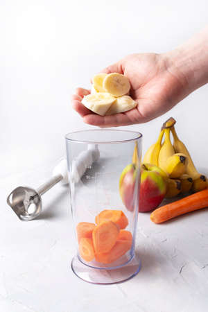 Step by step cooking smoothies. On a light background lies a blender, an apple, a banana, carrots and a measuring cup. A male hand puts a sliced banana in a glass. The concept of healthy nutritionの写真素材