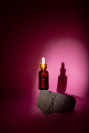 Cosmetic bottle with a pipette stands on a gray stone on a pink background. Directional light falls on the jarの写真素材