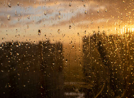 Close-up shot of raindrops on dirty glass. In the background in blur the silhouettes of city houses and the morning rising sunの写真素材