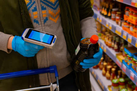 A customer wearing medical protective gloves holds a barcode scanner and scans the product. Cropped shot, no face visible. Self-service in the storeの写真素材