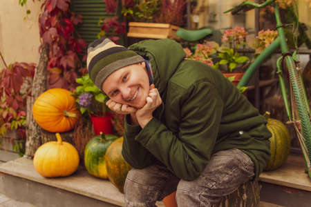 A young man sits on a stump against the backdrop of a shop decorated with pumpkins and flowers and smiles looking into the frame. Autumn warm colorsの写真素材