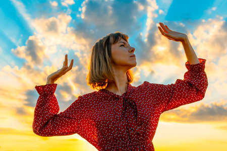 Young beautiful woman in a red dress against a blue sky with bright beams of the sun. The girl elegantly spread her arms in different directionsの写真素材
