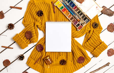 Flat lay of sketchbook, paints with brushes, yellow sweater, leaves, cinnamon sticks and pine cones on a white wooden background. Autumn composition with place for textの写真素材