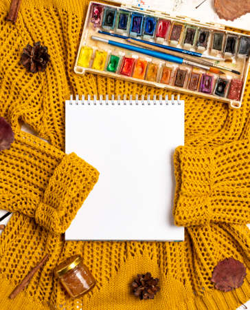 Flat lay of sketchbook, paints with brushes, yellow sweater, leaves, cinnamon sticks and pine cones on a white wooden background. Autumn composition with place for textの写真素材