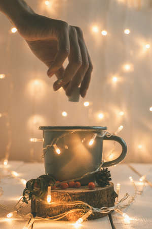 A female hand on the background of Christmas New Year's garlands puts sugar in a mug with hot sea buckthorn tea. Close-upの写真素材