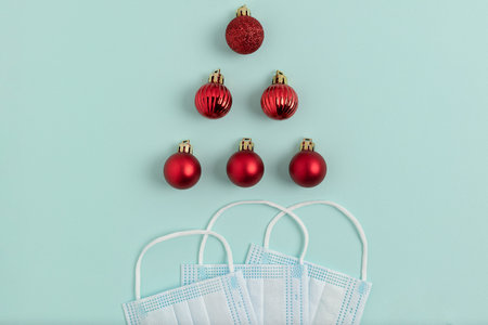 Three medical masks are fanned out and several red Christmas balls lie in the shape of a Christmas...