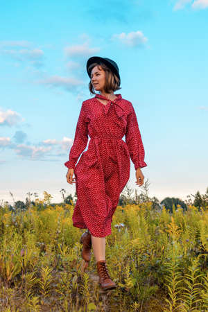 A young woman in a red dress and black hat walks in the field on a warm sunny dayの写真素材