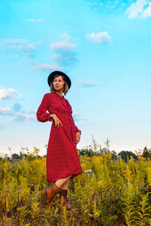 Young woman in red dress and black hat posing on the field on a warm sunny day.の写真素材