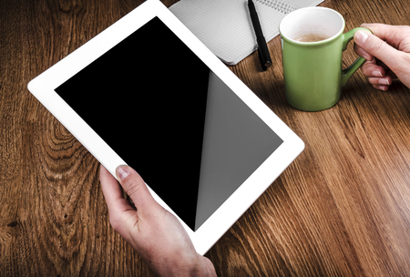Tablet with an empty screen in hands close to a pen and green cupの写真素材
