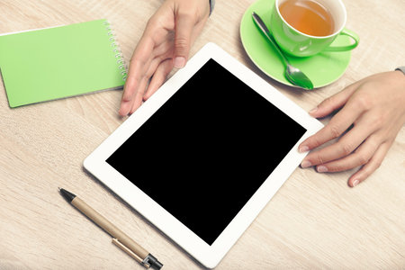 Tablet PC in the hands, a cup of tea and notebook on the desktop. Top view. Low contrast toned...