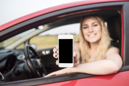 Happy blonde woman showing smartphone out the window of a car. Focus on mobile phone.の写真素材