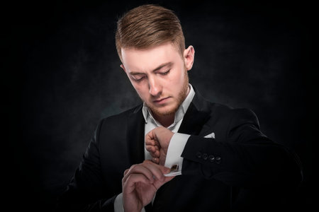 Elegant young fashion man looking at his cufflinks over dark background.