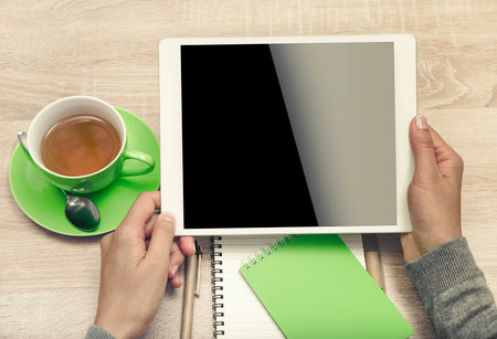 Tablet PC in the hands, a cup of tea and notebook on the desktop. Top view.の写真素材