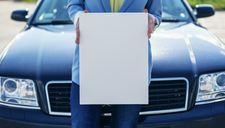 Woman standing by car holding a white blank poster.の写真素材