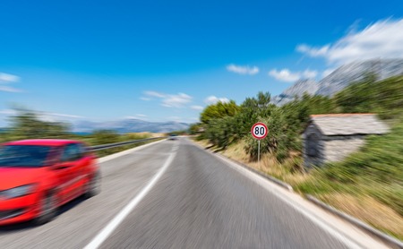 Road with a warning sign outside the city and a red car in motion.の写真素材