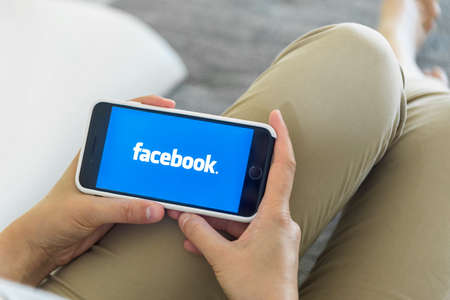 Woman Hands using iPhone with Facebook homepage on screen. Facebook the biggest social network website.のeditorial素材