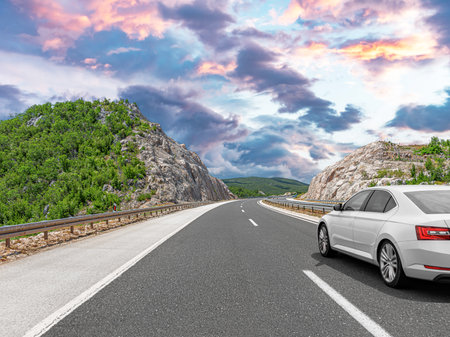 A white car drives along the highway against the backdrop of mountains on a sunny day.の写真素材