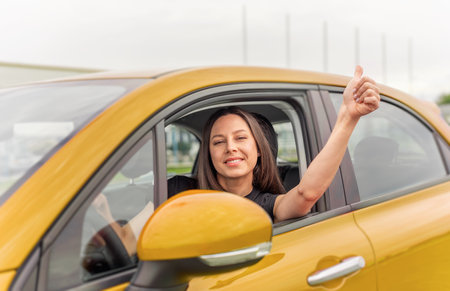 A young woman with bright smile sits in the drivers seat of a yellow car, giving a thumbs-up gestureの写真素材