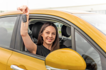 Woman with bright smile sits in the drivers seat of a yellow car, happily holding up a set of car...