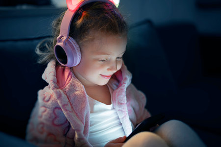 A young girl wearing pink glowing cat-ear headphones and a cozy fleece jacket is happily engaged with a mobile deviceの写真素材