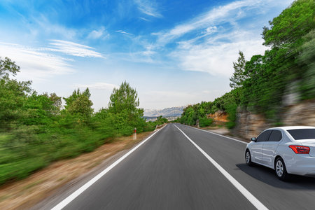 A white sedan driving on a blurred highway flanked by lush greenery under a partly cloudy blue sky