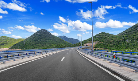 A modern highway with blue guardrails leads toward twin mountain tunnels surrounded by lush green hillsの写真素材