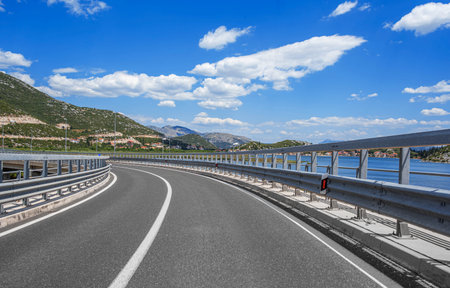 Scenic coastal highway with a curve and guardrails overlooking mountains, blue sky, and water