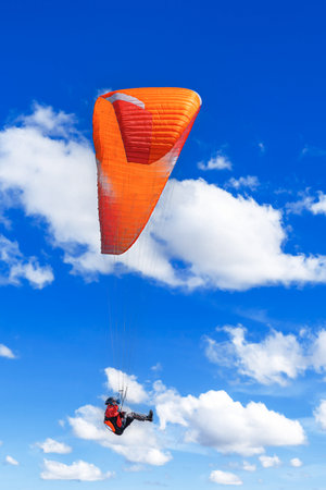 A lone paraglider with an orange wing soars high above the ground against a brilliant blue skyの写真素材