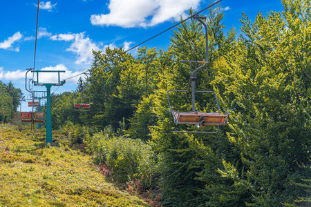 An old, rusty double chairlift ascends a grassy, sunlit ski slope surrounded by vibrant green deciduous treesの写真素材