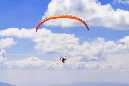 A paraglider with an orange canopy is silhouetted against a bright blue skyの写真素材