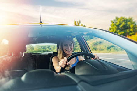 A happy woman with blonde hair smiles while driving her car on a sunny road during the golden hour.