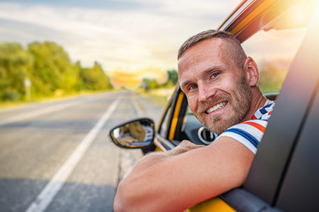 A happy man smiles at the camera while leaning out of the window of a yellow car on a sunny highway...