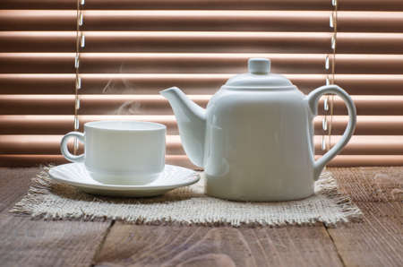 tea cup with teapot on old wooden table against the background of blindsの写真素材