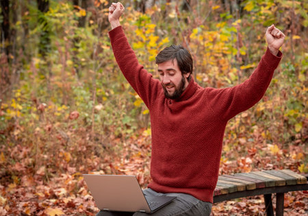 Man with cup of tea working freelance on laptop sitting outdoors on the park bench in the autumn...