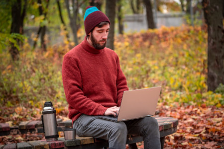 Man with cup of tea working freelance on laptop sitting outdoors on the park bench in the autumn...
