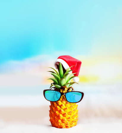 Attractive pineapple in stylish mirrored sunglasses on sand against sea water. Wearing christmas hat. Christmas and new year concept on a tropical beach.の写真素材