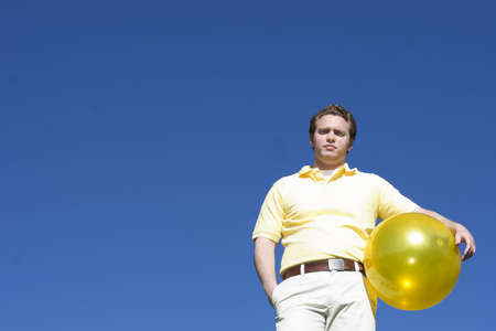 Man in a yellow shirt holding a yellow ball under a blue sky with an unhappy look on his faceの写真素材