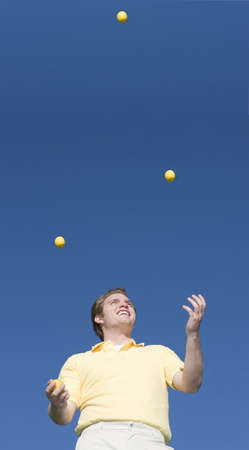 Young man with a smile juggles yellow balls in the air under a blue skyの写真素材