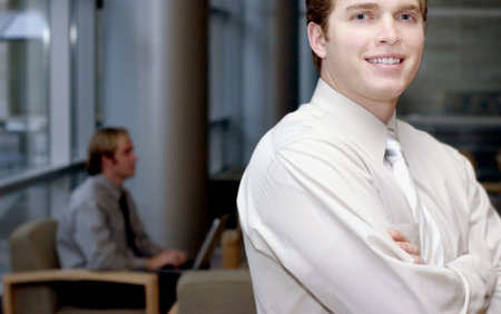 Young business man smiling looking forward with another business man in the background typing on...