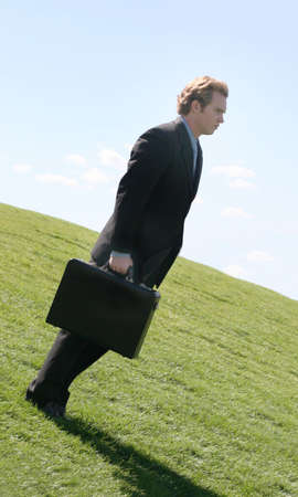 Business man in black suit and blue shirt holding briefcase as he is looking downwards on a green grass hill against the blue skyの写真素材