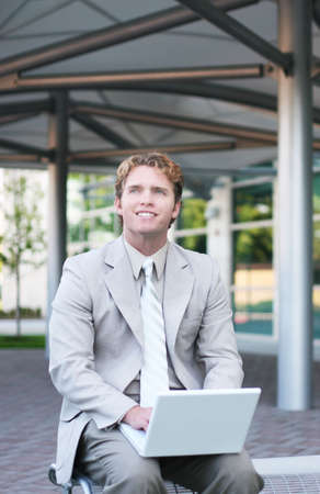 Business man in tan suit is sitting on park bench typing his white laptop and smilingの写真素材
