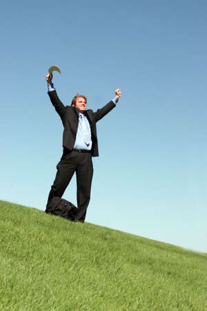 Businessman in black suit, blue shirt, and blue tie is holding his hands up in celebration as he stands in the green grass, against the blue skyの写真素材