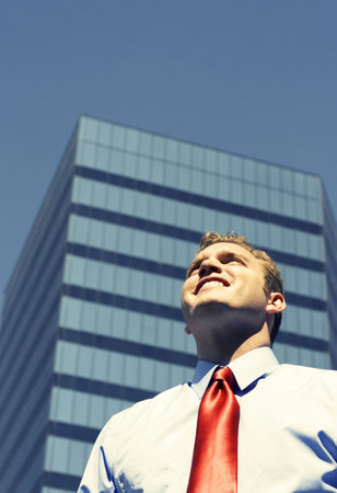 Business man with blue shirt and red tie is smiling in front of large, blue business buildingの写真素材