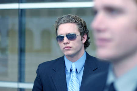 Business man in blue shirt and blue tie is wearing sunglasses with blurred shot of another business man in foregroundの写真素材