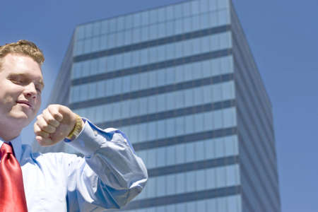 Businessman standing in front of an office building with his arm raised checking his gold watch for the timeの写真素材