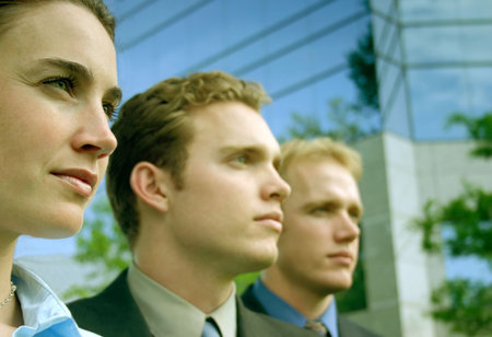 Three business people standing in a row standing in front of a blue business building all looking in...