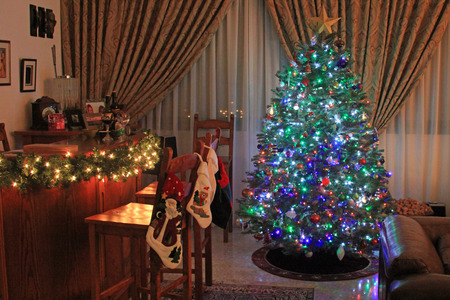 Festive Christmas tree decorated with multicolored lights and baubles next to a bar with pine garland and white lights and Christmas stockingsのeditorial素材