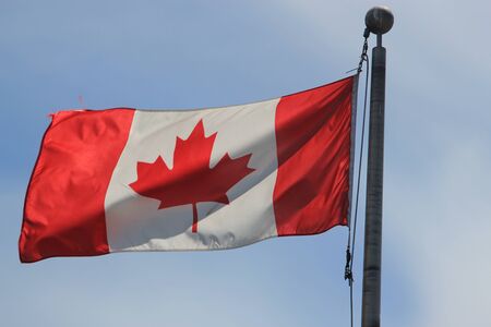 Canadian flag blowing to the left, with the complete flag and maple leaf on display, on a black flagpole against a blue sky backgroundの写真素材