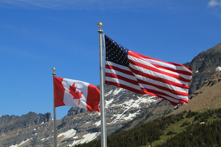 American and Canadian flags, showing the full stars and stripes and maple leaf respectively, on silver flagpoles blowing in the wind against a backdrop of snow covered mountains and forest treesの写真素材