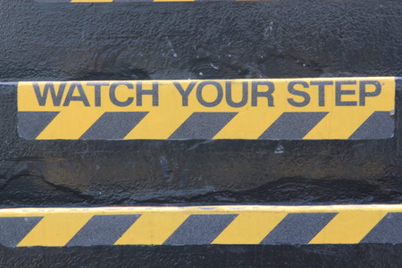 Yellow and black warning sign on stairs stating "Watch your Step" signifying a potential trip hazard, steep stairs and care to be taken when going up or down stairsの写真素材